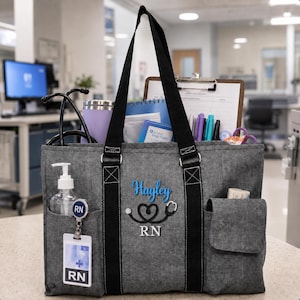 May include: A charcoal gray tote bag with black straps, embroidered with the name Hayley in blue, a black stethoscope heart icon, and the letters RN in white. The bag contains a stethoscope, a clipboard, pens, and a hand sanitizer bottle.