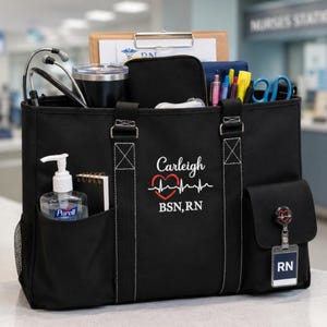 May include: A black canvas tote bag featuring white stitching, two handles, and exterior pockets. The front is embroidered with the name Carleigh, a red heart with an EKG line, and the text BSN, RN. It contains a stethoscope, hand sanitizer, pens, and a badge reel with an RN ID tag.