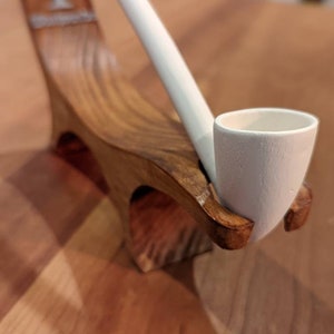 Może przedstawiać: Biały ceramiczny fajka spoczywa na drewnianym stojaku. Fajka ma długą, smukłą rurkę i zaokrągloną główkę. Stojak jest wykonany z ciemnego drewna i ma rzeźbiony wzór drzewa. Fajka i stojak są na drewnianej powierzchni.