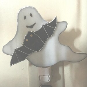 May include: A white stained glass ghost nightlight with a black bat on its back. The nightlight is plugged into an outlet.