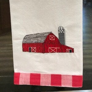 May include: A white kitchen towel with a red and white gingham border. The towel features an embroidered design of a red barn with a silo.