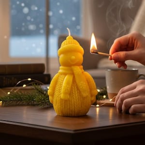 Könnte beinhalten: Eine gelbe Schneemann-Kerze mit Hut und Schal steht auf einem Holztisch. Ein brennendes Streichholz wird in die Nähe des Dochtes gehalten, Rauch steigt auf. Die Szene spielt sich in einem Innenraum ab, mit Büchern und einem Fenster im Hintergrund.