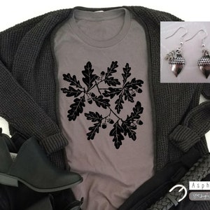 Puede incluir: Una camiseta gris con una silueta negra de hojas de roble y bellotas. La camiseta se combina con un c&aacute;rdigan negro, unos vaqueros negros y unas botas negras de tobillo. La imagen tambi&eacute;n incluye un par de pendientes de plata en forma de bellota.