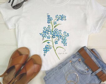 May include: White t-shirt with a watercolor illustration of blue forget-me-not flowers. The shirt is paired with blue jeans, brown sandals, sunglasses, and gold hoop earrings. The text "Forget Me Not" is visible.