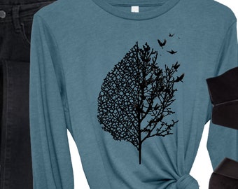 Könnte beinhalten: Ein langärmeliges Shirt in Heather Deep Teal mit einem schwarzen Grafikdruck eines Baumes, der auf der linken Seite in ein detailliertes Blattskelett übergeht und auf der rechten Seite kahle Äste mit wegfliegenden Vögeln zeigt, kombiniert mit schwarzer Jeans und Stiefeln.