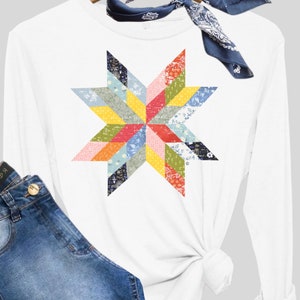 Pode incluir: Camiseta branca de manga comprida com um design de estrela de patchwork colorido. A estrela é feita de quadrados de tecido de diferentes cores com padrões florais. A camiseta é mostrada com um par de calças de ganga azuis e uma bandana azul.