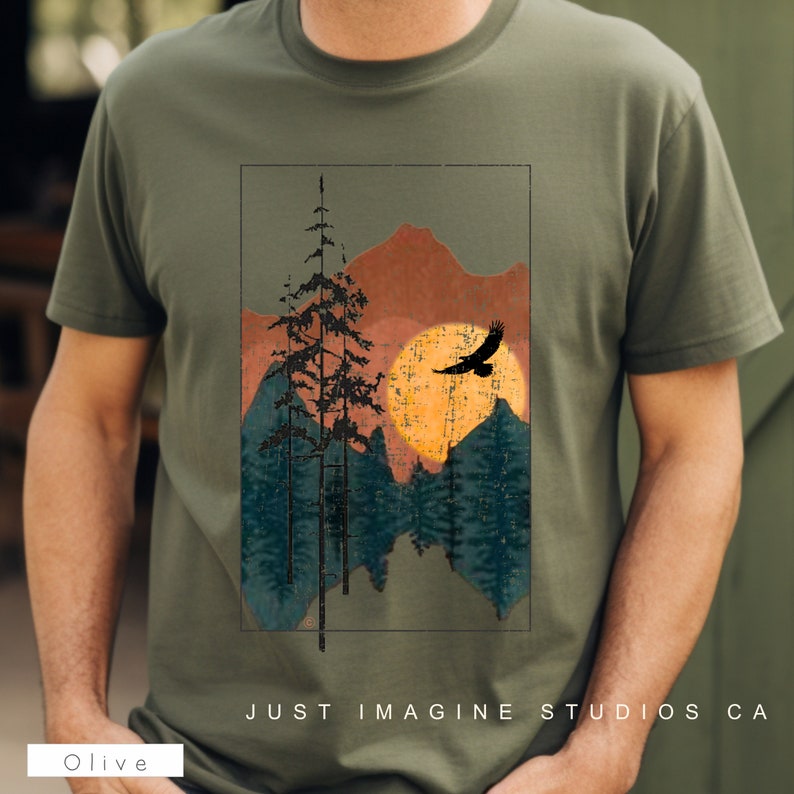 May include: Olive green t-shirt with a vintage-style graphic of a mountain range, a sun, and a bird flying over the mountains. The text "JUST IMAGINE STUDIOS CA" is printed on the bottom of the shirt.