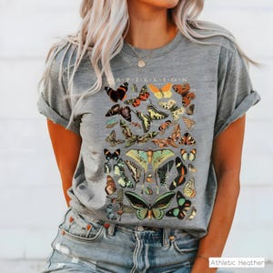 May include: Gray t-shirt with a colorful butterfly graphic. The design features various butterfly species in shades of orange, yellow, green, and black, with the word "APILLION" at the top. The shirt is made of a soft material.