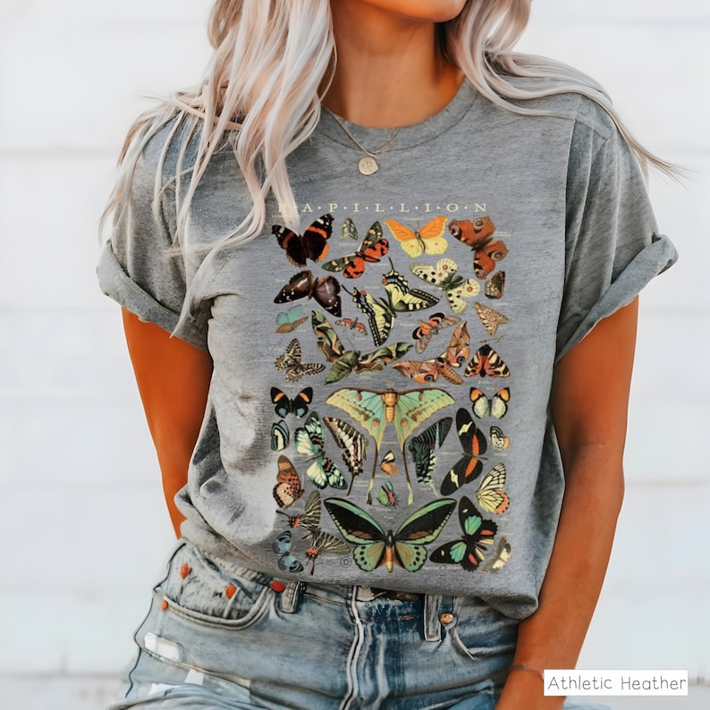 May include: Gray t-shirt with a colorful butterfly graphic. The design features various butterfly species in shades of orange, yellow, green, and black, with the word "APILLION" at the top. The shirt is made of a soft material.