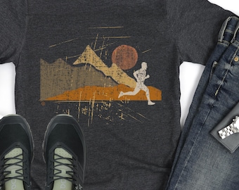 May include: Dark grey t-shirt featuring a mountain and sun graphic with a runner silhouette. The shirt is paired with blue jeans and black running shoes. A watch and wallet are also visible.