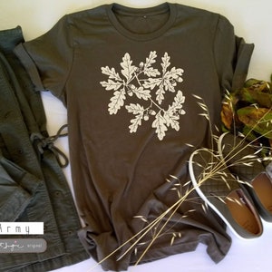 Puede incluir: Una camiseta verde oscuro con un estampado blanco de hojas de roble y bellotas. El texto "Army" est&aacute; impreso en la esquina inferior izquierda de la camiseta. La camiseta est&aacute; colocada sobre una superficie blanca con una chaqueta verde y una rama de hierba seca.