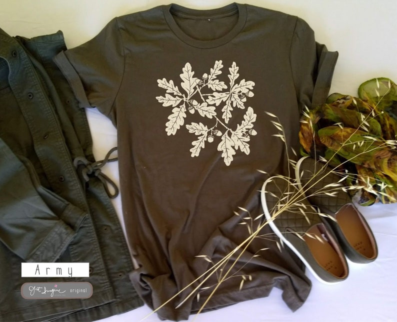 Puede incluir: Una camiseta verde oscuro con un estampado blanco de hojas de roble y bellotas. El texto "Army" est&aacute; impreso en la esquina inferior izquierda de la camiseta. La camiseta est&aacute; colocada sobre una superficie blanca con una chaqueta verde y una rama de hierba seca.