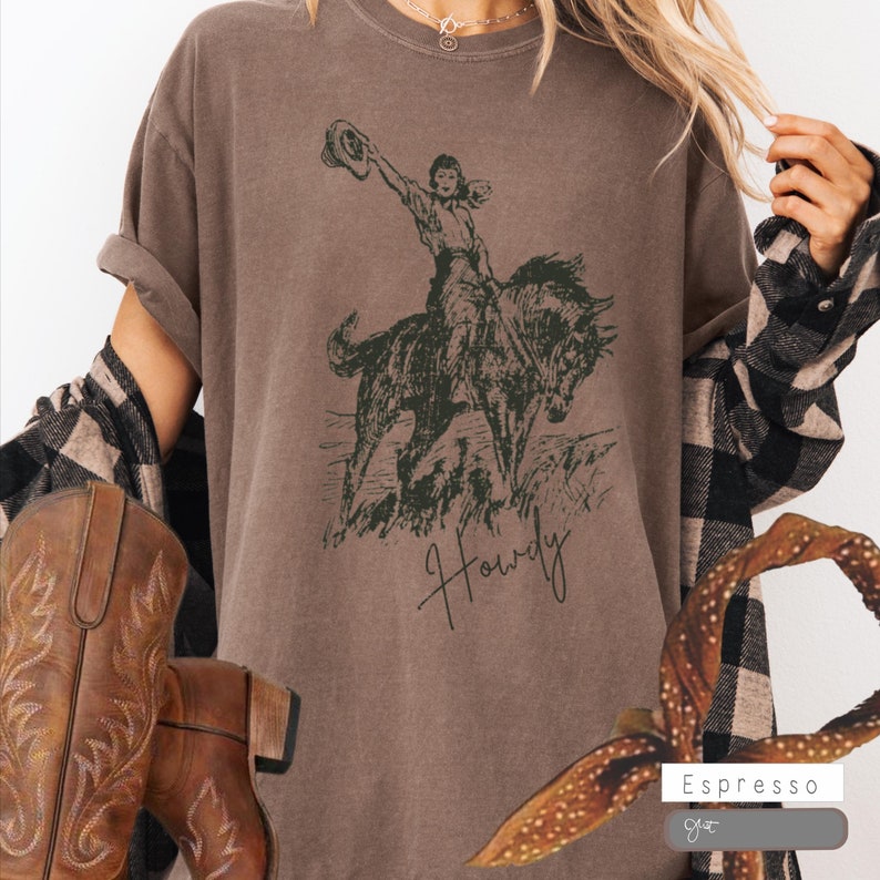 May include: A brown t-shirt with a vintage illustration of a woman riding a horse. The text "Howdy" is written in cursive below the image. The t-shirt is available in the color Espresso.