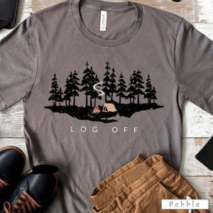 Puede incluir: Camiseta gris con un gráfico negro de una escena de bosque, una fogata y una tienda de campaña. El texto "LOG OFF" está impreso debajo. La camiseta está hecha de un material suave.