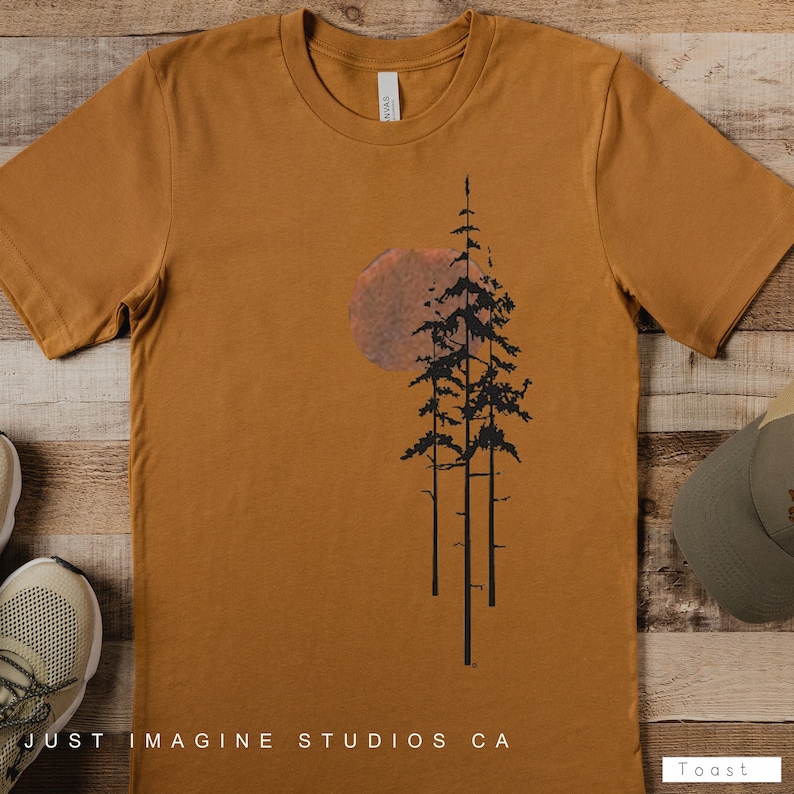 May include: A mustard yellow t-shirt with a black silhouette of three pine trees and a large orange sun behind them. The text "Just Imagine Studios CA" is printed at the bottom of the shirt.