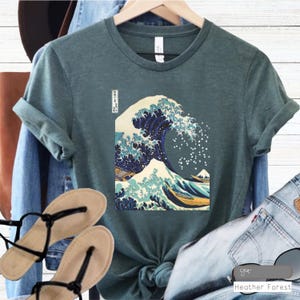 Pode incluir: Uma camiseta azul-esverdeada com uma estampa gráfica da Grande Onda de Kanagawa, uma famosa gravura japonesa em madeira de Hokusai. A impressão é em preto, azul, branco e amarelo.