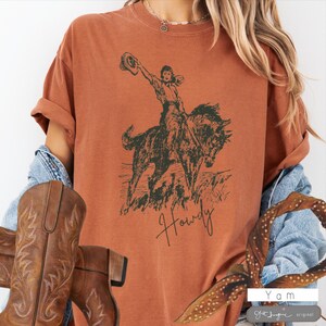 May include: A rust-colored t-shirt with a vintage illustration of a woman riding a horse. The text "Howdy" is printed below the image.