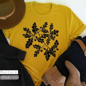 Puede incluir: Una camiseta amarilla mostaza con una silueta negra de hojas de roble y bellotas. El texto "Mustard" y "Get Imagine Original" est&aacute; impreso en la camiseta.