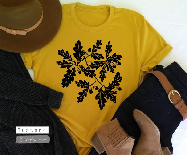 Puede incluir: Una camiseta amarilla mostaza con una silueta negra de hojas de roble y bellotas. El texto "Mustard" y "Get Imagine Original" est&aacute; impreso en la camiseta.