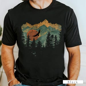 May include: Black t-shirt featuring a vintage-style graphic of a mountain range, a setting sun, and a flying bird. The design incorporates shades of orange, green, and brown, evoking a nature-inspired theme. The word "Black" is printed at the bottom right.