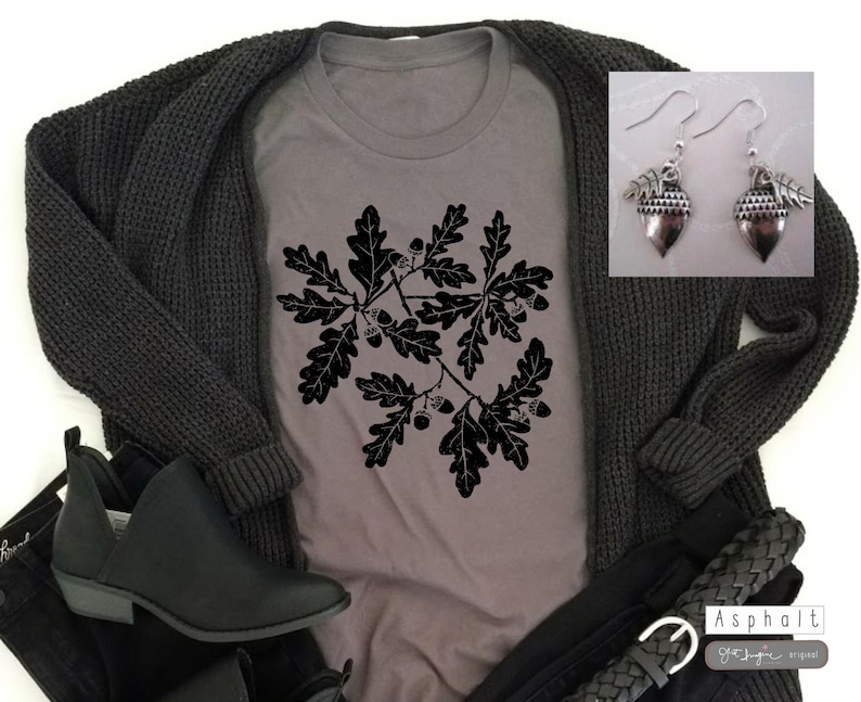 Puede incluir: Una camiseta gris con una silueta negra de hojas de roble y bellotas. La camiseta se combina con un c&aacute;rdigan negro, unos vaqueros negros y unas botas negras de tobillo. La imagen tambi&eacute;n incluye un par de pendientes de plata en forma de bellota.