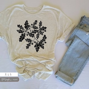 Puede incluir: Una camiseta beige claro con un gr&aacute;fico negro de hojas de roble y bellotas. La camiseta est&aacute; doblada y colocada sobre una superficie blanca junto a un par de jeans azul claro.