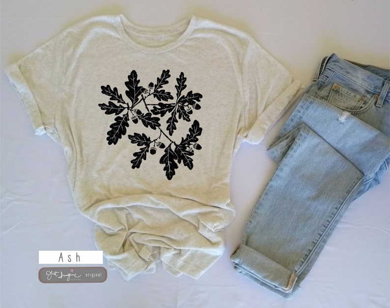 Puede incluir: Una camiseta beige claro con un gr&aacute;fico negro de hojas de roble y bellotas. La camiseta est&aacute; doblada y colocada sobre una superficie blanca junto a un par de jeans azul claro.