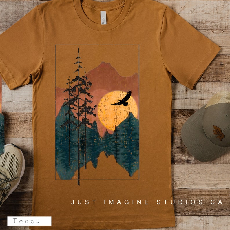 May include: A brown t-shirt with a graphic design of a sunset over a mountain range with trees and a bird flying in the sky. The text "JUST IMAGINE STUDIOS CA" is printed on the bottom of the shirt.