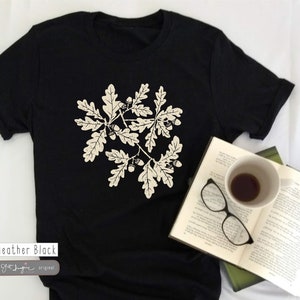 Puede incluir: Una camiseta negra con un estampado blanco de hojas de roble y bellotas. El texto "Heather Black" y "Oft Imagine original" est&aacute; impreso en la parte inferior de la camiseta.