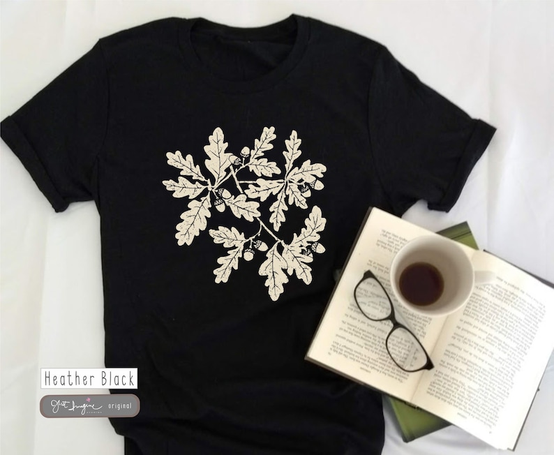 Puede incluir: Una camiseta negra con un estampado blanco de hojas de roble y bellotas. El texto "Heather Black" y "Oft Imagine original" est&aacute; impreso en la parte inferior de la camiseta.