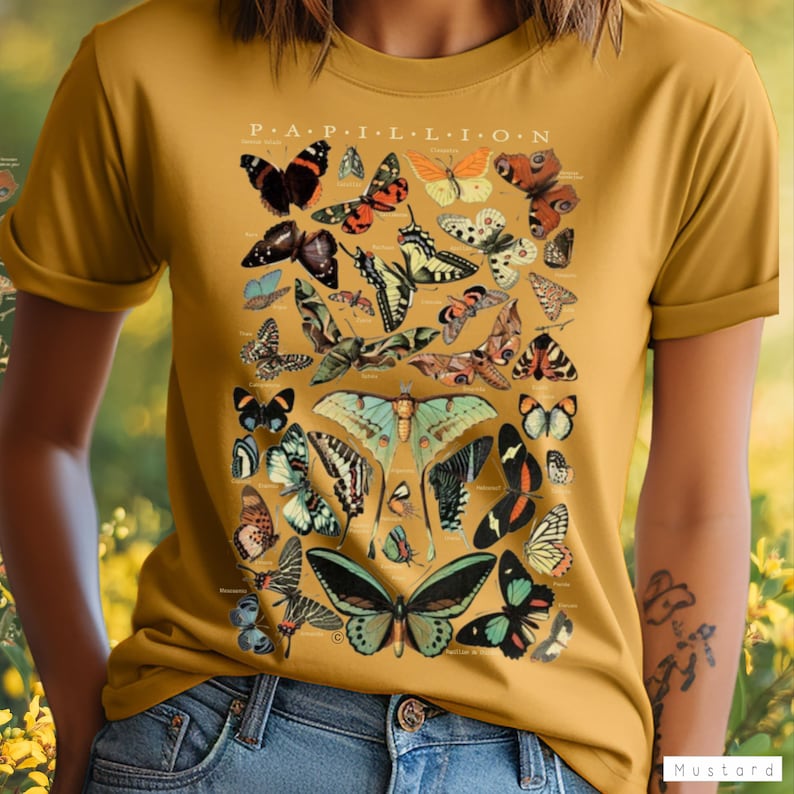 May include: Mustard yellow t-shirt featuring a detailed illustration of various butterflies. The design includes butterflies in shades of black, orange, green, and white, with the word "PAPILLION" at the top. The shirt is made of a soft material.