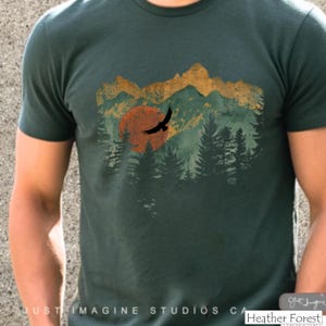 May include: Heather Forest green t-shirt featuring a nature-themed graphic. The design includes a sunset, mountains, and a flying bird silhouette. The text "Just Imagine Studios CA" is at the bottom of the shirt.