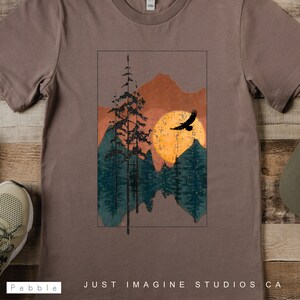 May include: A brown t-shirt with a vintage-style graphic of a mountain range with a sun and a bird flying over it. The graphic is in shades of orange, yellow, green, and blue. The text "JUST IMAGINE STUDIOS CA" is printed on the bottom of the shirt.