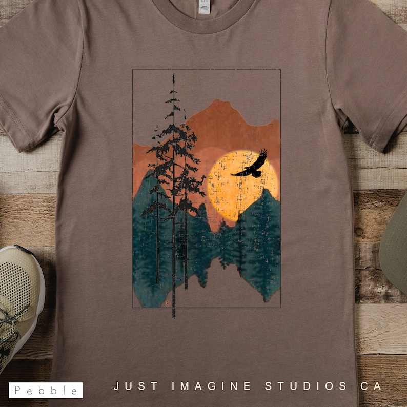 May include: A brown t-shirt with a vintage-style graphic of a mountain range with a sun and a bird flying over it. The graphic is in shades of orange, yellow, green, and blue. The text "JUST IMAGINE STUDIOS CA" is printed on the bottom of the shirt.