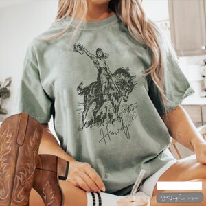 May include: Sage green t-shirt with a vintage illustration of a woman riding a horse. The text "Howdy" is printed below the image.
