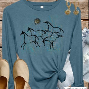 May include: A heather deep teal long-sleeve shirt featuring a black graphic print of three running horses and a spiral sun. The design includes distressed blue and tan paint splatter accents. The shirt is displayed with gold sun-shaped earrings.