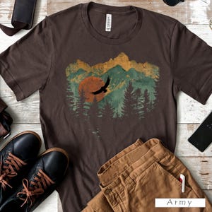 May include: A brown t-shirt featuring a nature-themed graphic. The design includes a sunset, mountains, and trees, with a silhouette of a flying bird. The shirt is paired with brown pants and black shoes, suggesting a casual style.
