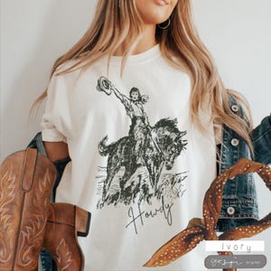 May include: A white t-shirt with a black and white illustration of a woman riding a horse. The text "Howdy" is written below the image.