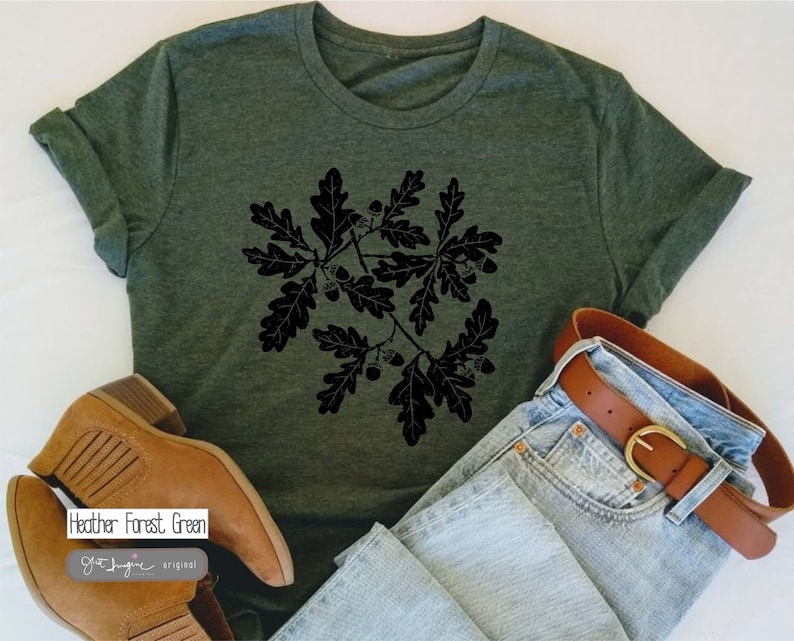 Puede incluir: Una camiseta verde bosque de color heather con un gr&aacute;fico negro de hojas de roble y bellotas. La camiseta se combina con jeans azules y botas marrones. El texto "Heather Forest Green" es visible en la camiseta.