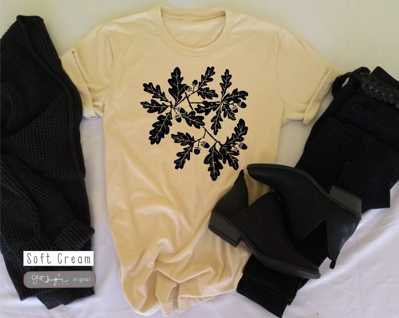 Puede incluir: Una camiseta de color crema suave con una silueta negra de una rama de roble con hojas y bellotas. El texto "Soft Cream" est&aacute; impreso en la esquina inferior izquierda de la camiseta. El texto "Gift Imagine Original" est&aacute; impreso debajo del texto "Soft Cream".