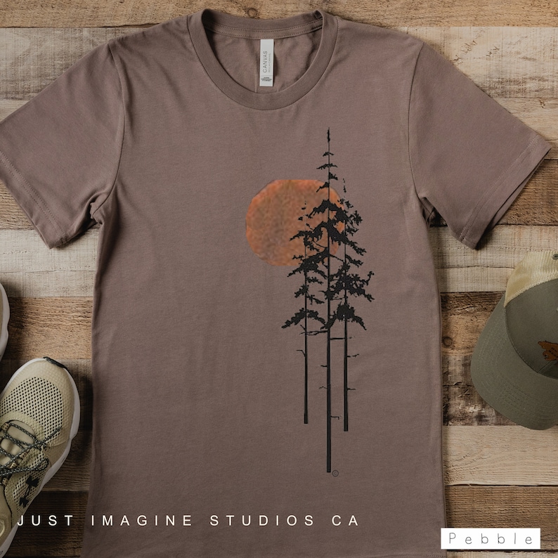 May include: A brown t-shirt with a graphic of three trees and a large orange sun. The text "JUST IMAGINE STUDIOS CA" is printed at the bottom of the shirt.