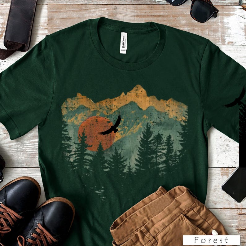 May include: Dark green t-shirt featuring a mountain and forest graphic with a setting sun and flying birds. The graphic has an aged, vintage look. The word "Forest" is printed at the bottom of the image.