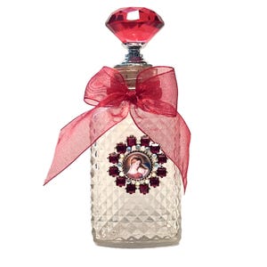 May include: A clear glass bottle with a diamond-shaped red topper, featuring a red sheer ribbon tied around the neck. The front of the bottle is decorated with a circular medallion containing a religious image surrounded by red and clear crystals.