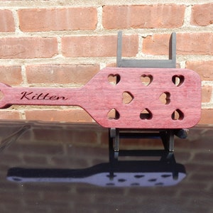 May include: A wooden paddle with a heart-shaped cutout design. The paddle is a deep reddish-purple color and has the word "Kitten" engraved on the handle.
