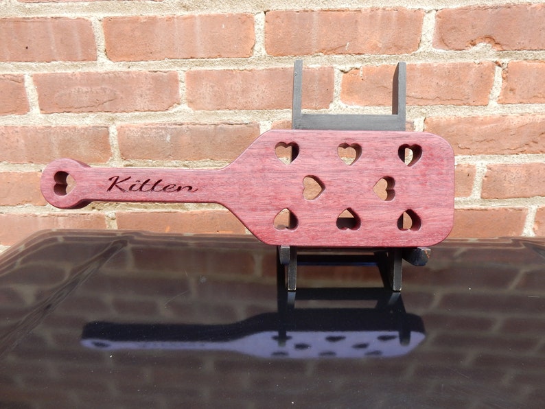 May include: A wooden paddle with a heart-shaped cutout design. The paddle is a deep reddish-purple color and has the word "Kitten" engraved on the handle.