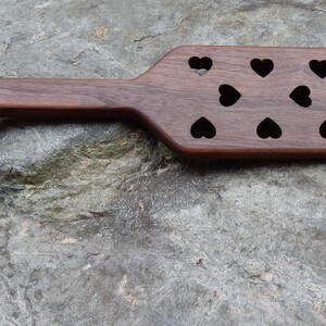 May include: A wooden paddle with a heart-shaped cutout at the top and multiple heart-shaped cutouts across the surface.