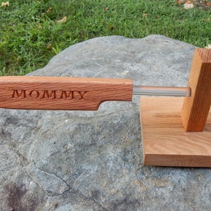 Puede incluir: Un utensilio de cocina de madera con la palabra "MOMMY" grabada en el mango, con un recorte en forma de corazón. La herramienta tiene una varilla transparente y está soportada por una base de madera. La madera tiene un tono marrón rojizo.