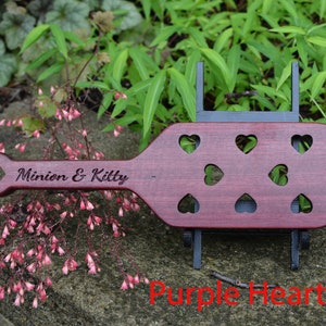 May include: A wooden paddle with a heart-shaped handle and multiple heart-shaped cutouts. The paddle is made of purple heart wood and is engraved with the text "Minion & Kitty".