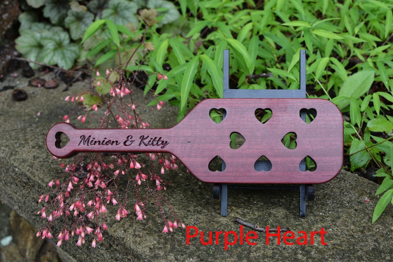 May include: A wooden paddle with a heart-shaped handle and multiple heart-shaped cutouts. The paddle is made of purple heart wood and is engraved with the text "Minion & Kitty".