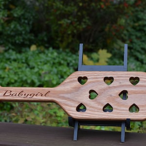 May include: A wooden paddle with a heart-shaped cutout at the end of the handle. The paddle has a carved design of hearts and the words "Babygirl" on the handle.